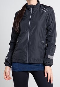 Chaqueta ligera negra con cierre de cremallera, cuello alto, detalles reflectantes y puños elásticos. El material es suave y resistente al agua.