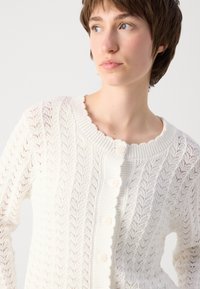Weiße Kabelstrick-Cardigan mit gewelltem Ausschnitt, Knopfschluss und offenem Strickmuster. Texturierter Stoff mit weichem Finish.