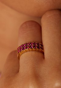 Gouden ring met een rij roze edelstenen langs de bovenkant, met een gestructureerde band. De edelstenen zijn rond en dicht bij elkaar gezet, wat een levendig uiterlijk creëert.