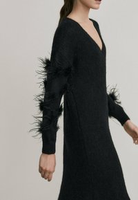 Black knitted dress with a V-neck and long sleeves. Features textured black feathers along the sleeve edges. Soft, form-fitting design.