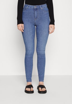 Højtaljede blå denim skinny jeans med en glat tekstur, fem lommer og knaplukning. Sammen med sorte flade sandaler.