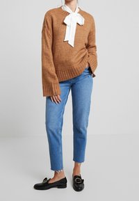 Persoon in een bruin gebreid sweater met witte strikdetails, lichtblauwe jeans met rafelrand en zwarte instap loafers met een gouden gesp.