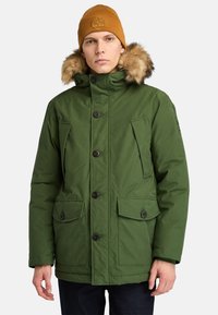 Groene geïsoleerde jas met een bontgevoerd capuchon, voorknopjes, twee zijzakken en een gestructureerde stof. Draagt een bruine gebreide beanie.