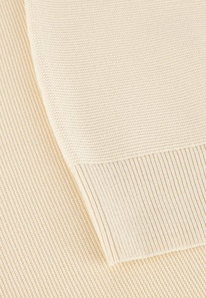Cremefarbene Strickstoff mit gerippter Textur. Nahaufnahme, die die gebogene Kante und die ausgeprägten Muster zeigt, die aus dem Nähen resultieren.