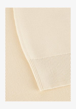 Material tricotat de culoare crem, cu textură canelată. Vedere detaliată care arată marginea curbată și modelele distincte rezultate din coasere.