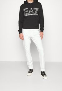 Hombre con sudadera negra de Emporio Armani, pantalones blancos y zapatillas negras de caña alta, de pie contra un fondo blanco.