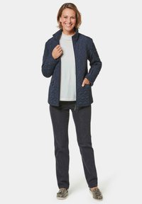 Marineblauer Steppjacke mit hohem Kragen, Reißverschlüssen und Seitentaschen, kombiniert mit einem hellen Hemd und dunkler Jeans. Die Schuhe haben ein Schlangenledermuster.