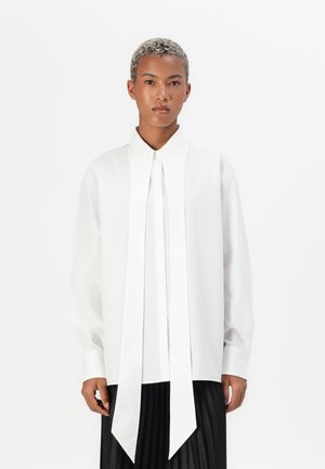 White long-sleeve shirt with a sharp collar and a front placket. Features an elongated tie detail at the neck. Smooth, structured fabric.