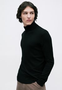 Jovem com cabelo escuro e ondulado, vestindo um suéter preto de gola alta e calças castanhas, olhando para o lado contra um fundo branco simples.