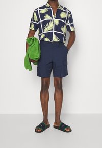 Navy shirt with short sleeves and a green leaf pattern, paired with navy cargo shorts, holding a green towel, and wearing black sandals.