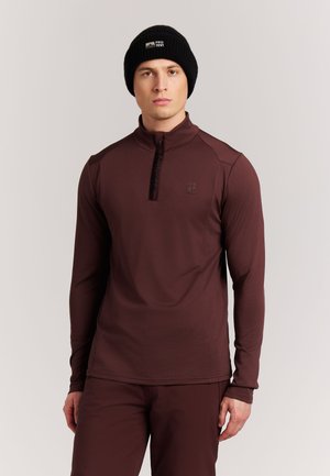 Man draagt een zwarte gebreide muts en een donkerbruine longsleeve met een kwart rits en bijpassende broek, staande tegen een effen achtergrond.