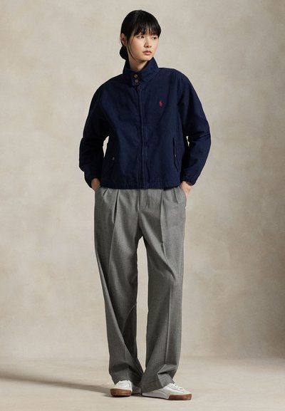 Tumši zila kokvilnas rāvējslēdzēja jaka ar augstu apkakli un logotipa akcentu. Kombinēta ar pelēkām kroku biksēm un baltām kedām.