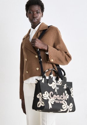Personne noire portant une veste croisée marron et un pantalon blanc, tenant un sac à main floral noir et blanc Coach New York.