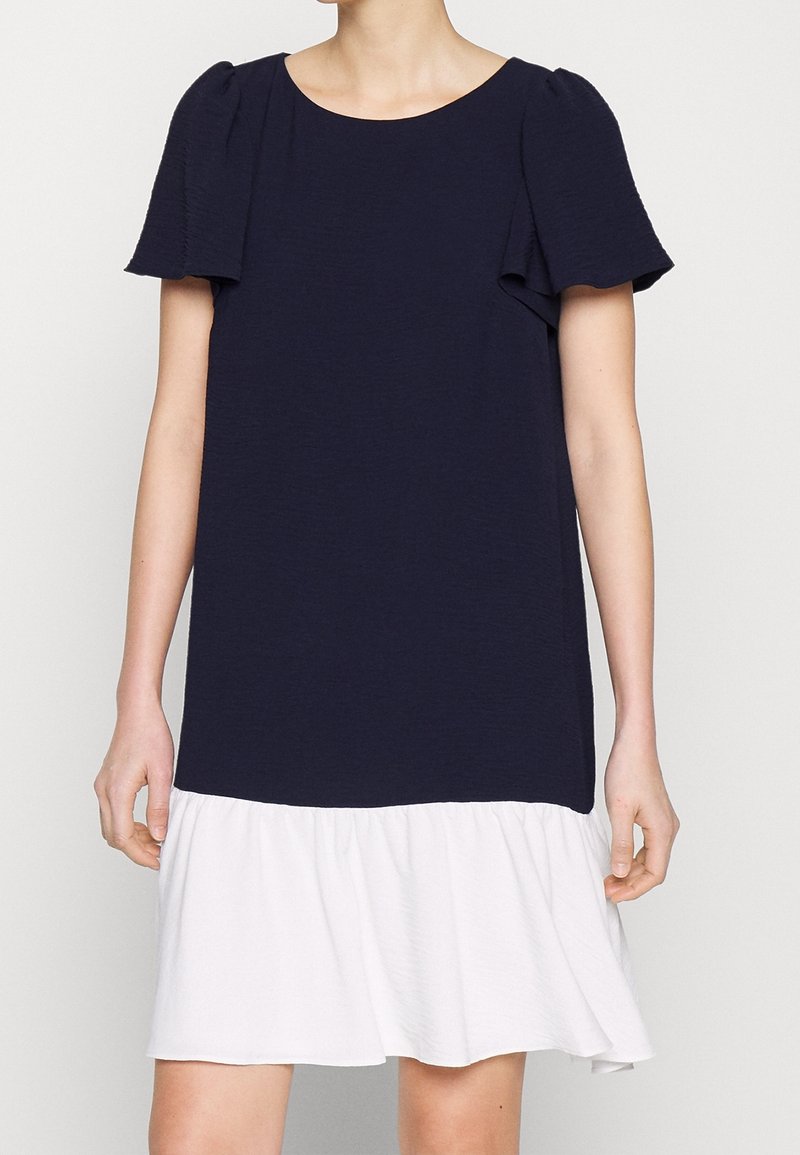 Robe bleu marine à manches courtes avec un col rond et un ourlet blanc à volants, portée par une personne se tenant devant un fond uni.