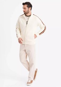 Veste zippée blanche à rayures latérales marron, associée à un pantalon beige clair et des baskets marron. Le tissu semble doux avec un design décontracté.