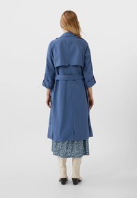 Niebieski płaszcz trencz z paskiem w talii, długimi rękawami i detalem klapy z tyłu, noszony na sukience w kwiatowy wzór oraz kremowych kozakach do kolan.