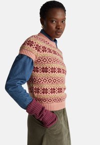 Růžový pletený svetr s květinovými a geometrickými vzory, doplněný džínovou košilí a texturovanými vínově červenými rukávy. Zelené manšestrové kalhoty.