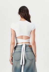 Valkoinen puuvillainen lyhythihainen crop-top, jossa on selkäaukko ja sidontanauhat. Yhdistetty korkeavyötäröisiin vaaleansinisiin farkkuihin.