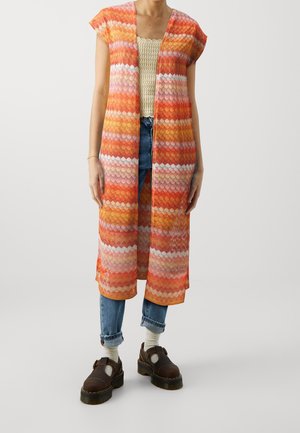 Kobieta ubrana w pomarańczowo-różowo-biały wzorzysty bezrękawnik na długi fason, kremowy top z szydełkowego materiału, podwinięte niebieskie dżinsy, białe skarpetki i brązowe buty na platformie.