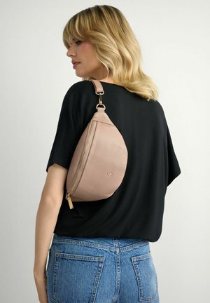 Blondynka w czarnej bluzce i niebieskich dżinsach, z beżową skórzaną torbą na ramię, na jednolitym tle.