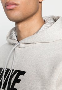 Svetle šedá mikina s kapucí a černým logem, která má texturované stříbrné lemování v kapuci, stahovací šňůrky a je vyrobena z měkké směsi bavlny.