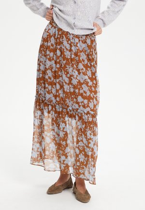 Kobieta w brązowej maxi spódnicy z białym kwiatowym wzorem, w połączeniu z jasnoszarym dzianinowym kardiganem i brązowymi zamszowymi balerinami.