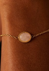Gouden kettingarmband met een ovale roze steen omgeven door ingewikkelde details, geplaatst tegen een textuur van huid.