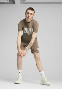 Conjunto deportivo marrón que incluye una camiseta de manga corta con un estampado brillante de "PUMA sports club" y pantalones cortos a juego, combinados con zapatillas blancas.
