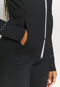 Personne portant une veste de sport noire avec une fermeture éclair blanche et des points réfléchissants, la main partiellement dans la poche latérale d’un pantalon noir.