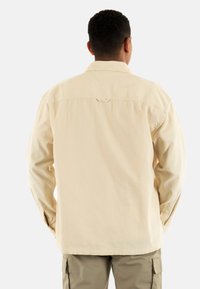 Camicia a maniche lunghe color beige chiaro con colletto a bottoni, tessuto testurizzato e dettagli sul yoke posteriore. Presenta maniche ampie con polsini a bottone.