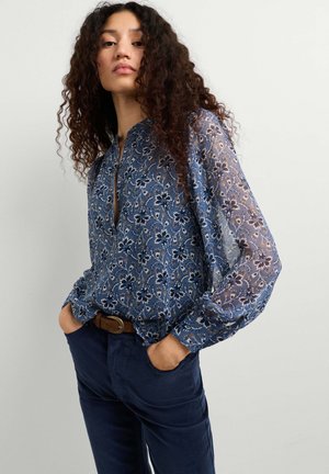 Frau mit lockigem Haar, die eine blaue, geblümte Langarmbluse und dunkelblaue Hose trägt, steht mit den Händen in den Taschen vor einfachem Hintergrund.