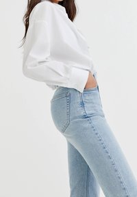 Camisa blanca de manga larga con cuello, combinada con jeans de tiro alto de color azul claro. La tela parece suave, con un estilo clásico de cinco bolsillos.