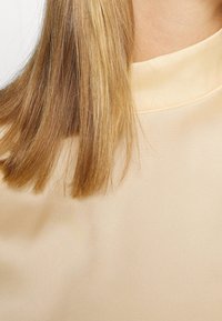 Haut en soie beige à col montant, texture lisse et coutures délicates, complété par de longs cheveux bruns et raides.