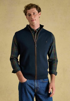 Giovane uomo che indossa un gilet blu navy con zip sopra una camicia a quadri verde e rossa con le maniche arrotolate e jeans blu, in piedi davanti a uno sfondo beige.