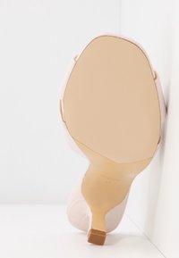 Vue de la semelle d'une chaussure à talons hauts rose pâle avec une semelle beige et un petit talon carré reposant contre une surface blanche.