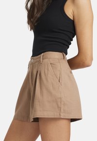 Femme portant un short beige taille haute sur mesure avec des plis à l'avant et un top noir sans manches côtelé, court à la taille.