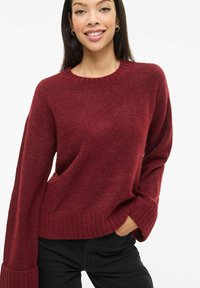 Burgundia punane kootud džemper pikkade, laiade varrukate ja soonikkoes mansettidega. Pehme tekstuur ja klassikalise ümmarguse kaelusega, sobitatud tume teksapükstega.