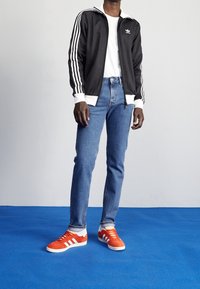 Person klädd i en svart Adidas träningsjacka, vit t-shirt, blå jeans och klar orange Adidas sneakers som står på en blå golv.