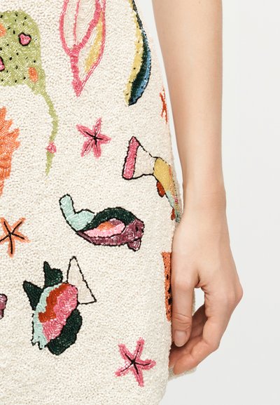 Κοντινό πλάνο ενός λευκού ενδύματος με πολύχρωμα μοτίβα από χάντρες σε σχήμα κοχυλιών, αστερία και κοραλλιών, δίπλα σε ένα χέρι που ακουμπάει.