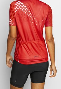Czerwona koszulka kolarska z krótkim rękawem z gradientowym wzorem serca, wykonana z oddychającego materiału. Posiada logo i trzy tylne kieszenie. Nosi się z czarnymi szortami.