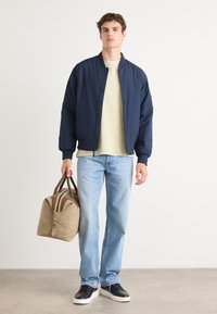 Veste de bombardier navy, t-shirt beige, jeans bleu clair et baskets noires ; portant un sac de sport beige avec des accents marron et une fermeture éclair.