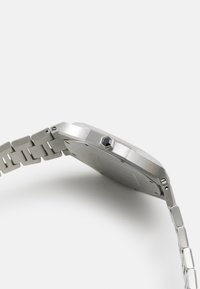 Montre en acier inoxydable argenté avec un boîtier octogonal, un bracelet texturé et une couronne hexagonale. Surface lisse et détails de finition polie.