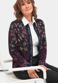 Navy vest met bloem- en paisleypatronen in rood, groen en blauw. Lichtgewicht gebreide stof met een getailleerd ontwerp. Witte overhemd met kraag eronder.