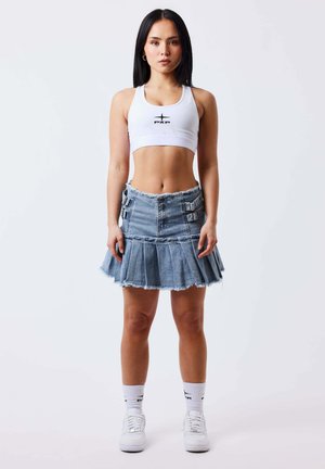 Giovane donna in piedi di fronte, indossa un reggiseno sportivo bianco, una gonna di jeans plissettata con fibbie, calzini bianchi e scarpe da ginnastica bianche su sfondo semplice.