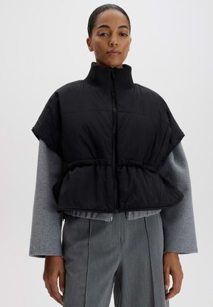 Frau, die eine schwarze, kurzärmelige, gesteppte Weste über einem grauen Langarmshirt und grauen, maßgeschneiderten Hosen vor einfarbigem Hintergrund trägt.