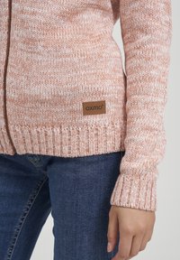 Närbild av en person som bär en rosa och vit stickad zip-tröja med en brun "Oxmo"-etikett och blå jeans, med handen avslappnad vid sidan.