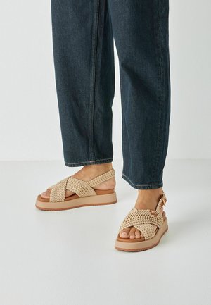 Beige geweven sandalen met gekruiste bandjes, lichtbruine zool, verstelbare enkelband en getextureerd bovenwerk. Gecombineerd met donkerblauwe spijkerbroek.