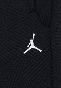 Čierna textúrovaná tkanina s geometrickým vzorom. Na boku sa nachádza bielo vyšívané logo Jumpman v dynamickej póze.
