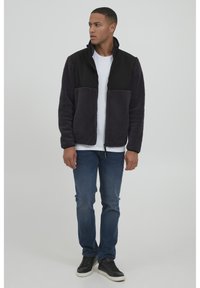 Fleecejakke med svart overpanel, grå kropp, glidelås og høy krage. Kombinert med blå jeans og svarte sneakers.