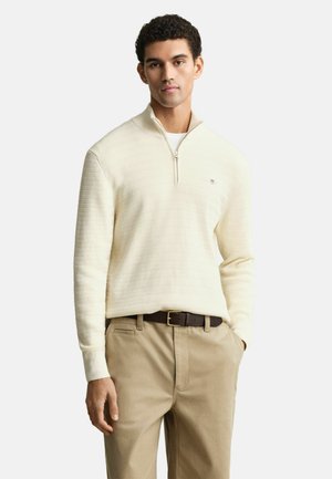 Cremfarbener Pullover mit Viertel-Reißverschluss und strukturiertem Strickdesign, über einem weißen Hemd getragen. Kombiniert mit beigen Hosen und einem braunen Gürtel.